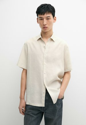 Ung mand iført en beige kortærmet skjorte og mørke jeans, stående med den ene hånd i lommen mod en ensfarvet baggrund.
