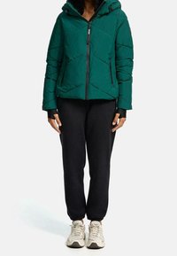 Giacca verde imbottita con cappuccio, design a cuciture diagonali, zip frontale completa e dettagli neri. Abbinata a pantaloni joggers neri e sneakers chiare.