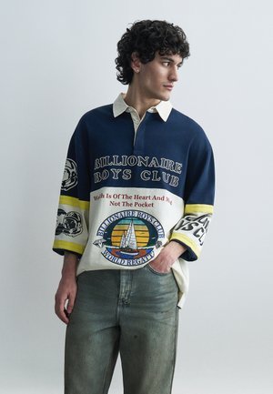 Młody mężczyzna o kręconych włosach w granatowo-kremowej koszulce rugby Billionaire Boys Club z logo żeglarstwa i zielonych dżinsach, patrzący w prawo.