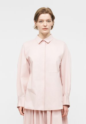 Kvinde med kort hår iført en lyserød skjorte med lange ærmer og knapper samt en matchende nederdel mod en ensfarvet hvid baggrund.