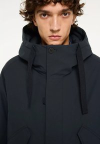 Zwarte capuchonjack met een gestructureerde afwerking, voorzien van een knoopsluiting, trekkoorden en hoekige voorzakken.