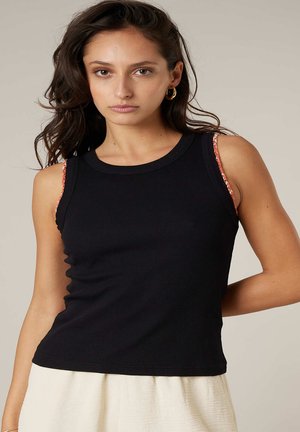 Jeune femme aux cheveux foncés portant un haut noir sans manches sur une sous-couche à motifs orange, associé à une jupe crème, fond neutre.