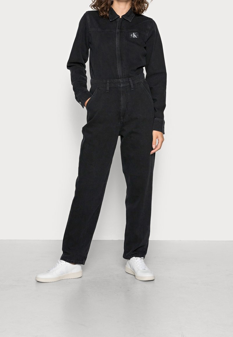 Personne portant une combinaison longue noire Calvin Klein avec col et baskets blanches, debout avec une main dans la poche.