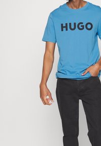 Jasnoniebieska koszulka bawełniana z okrągłym dekoltem i krótkimi rękawami, z wyrazem "HUGO" w pogrubionych czarnych literach na piersi.