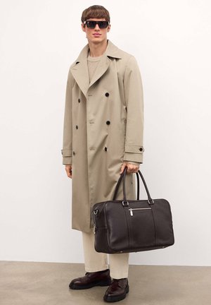Beige, dupla gombos trench coat, textúrált barna bőr táska felső fogantyúkkal és cipzárral, krém színű pulóver, krém színű nadrág, és sötét cipők.
