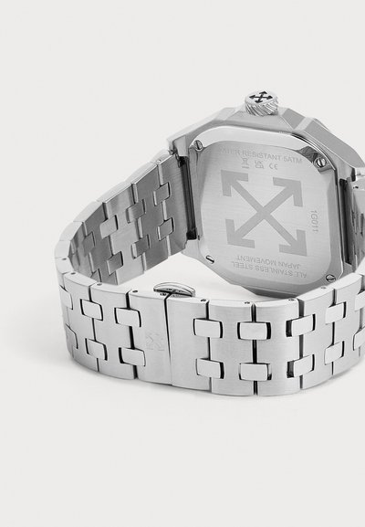 Montre-bracelet en acier inoxydable argenté avec bracelet lié, affichant le fond du boîtier gravé de flèches et des détails sur la résistance à l'eau.