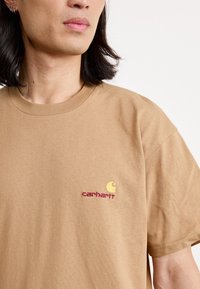 Tan cotton t-shirt featuring a round neckline and short sleeves, with a small embroidered logo in yellow and red on the chest.