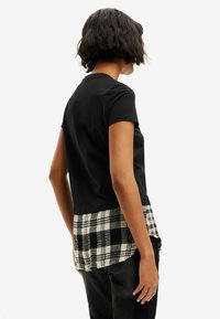Musta lyhythihainen t-paita, jossa on kerroksellinen helma mustavalkoisella ruutukuvioinnilla, esitetty takaa, sileällä pinnalla.