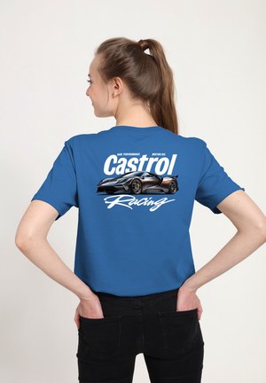 Frau mit Pferdeschwanz, die ein blaues T-Shirt trägt, auf dessen Rückseite ein schwarzes Sportauto und das weiße "Castrol Racing"-Logo zu sehen sind, Hände in den hinteren Taschen.