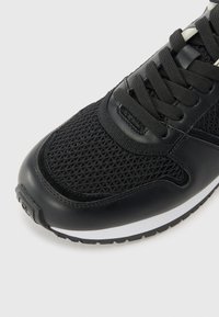 Black athletic shoe featuring a mesh upper and leather accents, with a textured surface and a white midsole. Prominent lacing system shown.