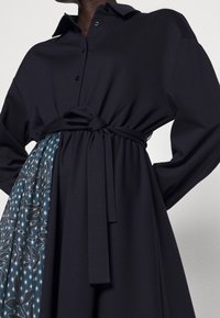 Abito nero a maniche lunghe con colletto e cintura, caratterizzato da un tessuto fantasioso in un design turchese e bianco su un lato.
