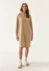 Vestido sudadera beige de tejido suave, con un ajuste relajado, capucha con cordón y puños de canalé. Combinado con zapatillas crema y calcetines claros.