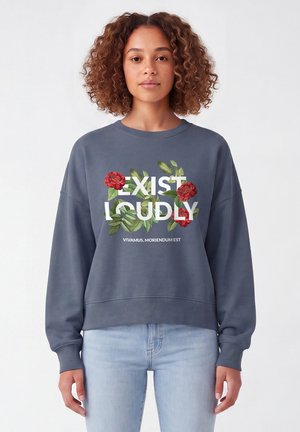 Frau mit lockigem Haar, die einen blauen Pullover mit roten Blumen und dem Text "EXIST LOUDLY" sowie kleinerem Text darunter trägt.