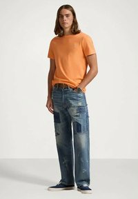 T-shirt de algodão laranja com gola redonda, combinada com jeans azuis desfeitos com detalhes em patchwork e corte descontraído.
