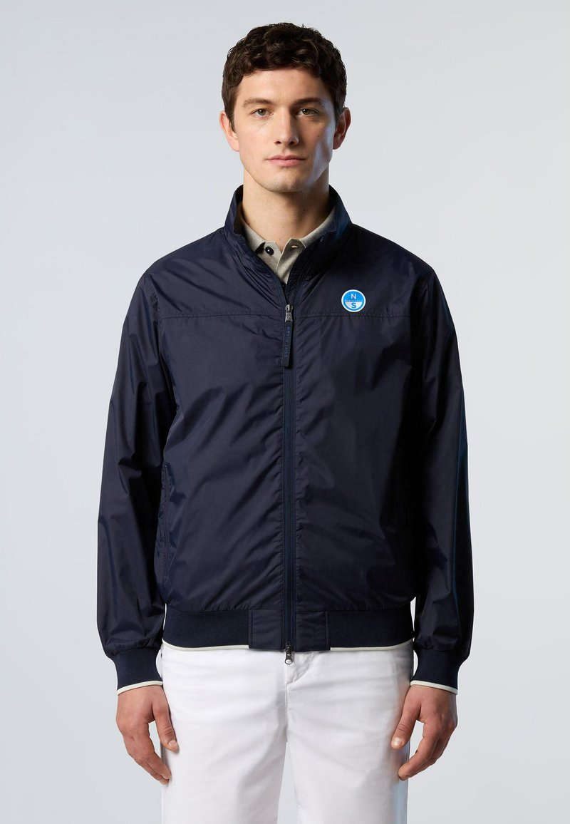 Giacca leggera zip-up blu navy con polsini e orlo elasticizzati, caratterizzata da un logo circolare sul petto e tessuto liscio e resistente all'acqua.