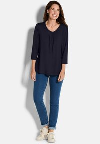 Marinefarbener Langarm-Top mit rundem Halsausschnitt und gefältelten Details, kombiniert mit blauen Jeans und cremefarbenen Sneakers. Lässiger, entspannter Schnitt.