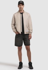 Jachetă bomber bej cu mâneci elastice, tricou negru dedesubt, pantaloni scurți din denim închisi la culoare și adidași negri, toate pe un fundal neutru.