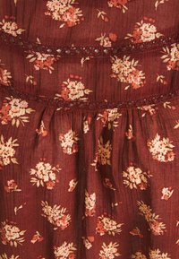 Tissu fluide bordeaux avec de petits motifs floraux en crème et rose. Présente une délicate bordure en dentelle entre des rangées horizontales de tissu.