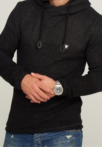 Hoodie noir en tricot avec cordons. Tissu texturé et petit logo sur la poitrine. Porté avec une montre-bracelet argentée.