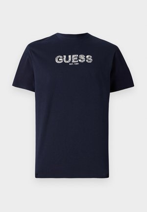Mornariča bombažna majica s kratkimi rokavi, na kateri je bela stilizirana beseda "GUESS" ter "EST. 1981" spodaj. Preprost, sproščen dizajn.