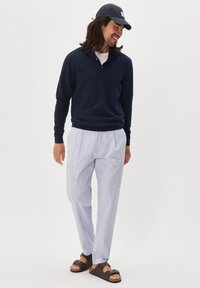 Marinefarbener Pullover mit Kragen, hellgestreifte Hose in Blau und Weiß sowie dunkle Sandalen mit Schnallen-Detail. Mann steht in einer entspannten Pose.
