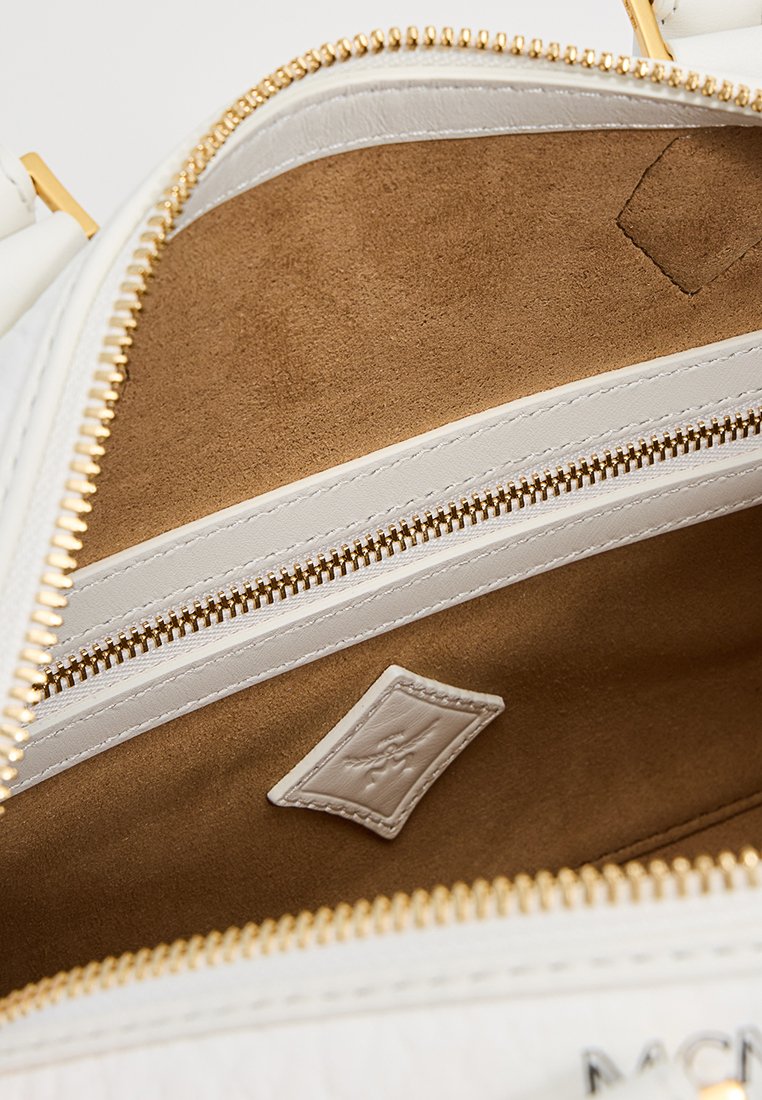 Weißes Lederhandtascheninnere mit beigem Wildlederfutter, ausgestattet mit einer Reißverschlusstasche und einem flachen Etikett mit geprägtem Logo. Goldfarbene Hardware-Akzente.