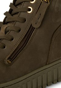 Botte de cheville en suede vert olive avec une fermeture éclair sur le côté, des lacets ronds, des œillets dorés, et une semelle en caoutchouc texturée avec des rainures horizontales.