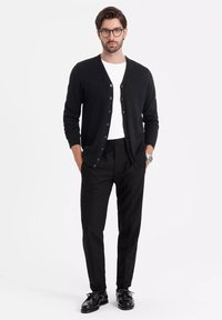 Svart cardigan med knappar, bärs över en vit t-shirt, ihop med svarta byxor och svarta snörskor. Minimalistisk stil, inga mönster.