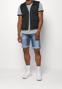 Svart tygväst med flera fickor, bärs över en randig t-shirt. Matchas med ljusblå denimshorts och vita sneakers.