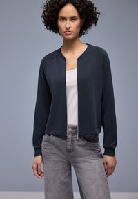 Blouson aviateur bleu marine foncé avec fermeture éclair, poignets côtelés et coupe décontractée, associé à un jean gris clair et un t-shirt blanc en dessous.