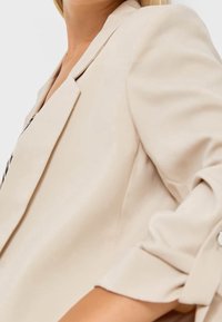 Blazer beige avec une coupe sur mesure, des revers structurés et des manches retroussées. Tissu lisse avec un léger éclat et des boutons en accent.