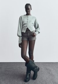 Sivý skrátený kardigán s gombíkmi, kombinovaný so sivými šortkami. Model má na sebe čierne čižmy po kolená s kovovými doplnkami. Neutrálny pozadie.