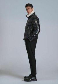 Black puffer jacket with a glossy finish and a white shearling collar. Features zippered pockets and a slim fit design. Black trousers and shoes complete the look.