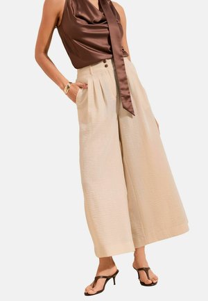 Kvinde iført beige vide bukser med kort længde, brun satin ærmeløs top med bindedetalje, guldbøjlearmbånd og brune hælede sandaler.