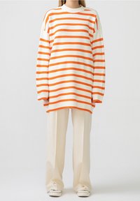 Gestreifter Pullover in Weiß und Orange mit Rundhalsausschnitt und weiter Passform. Kombiniert mit cremefarbenen Hosen mit weitem Bein und weißen Sneakers.