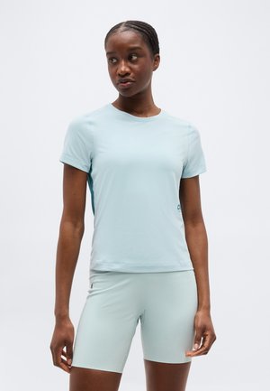 Mujer con el cabello trenzado, vestida con una camisa de manga corta azul claro y pantalones cortos hasta la rodilla a juego, de pie frente a un fondo liso.