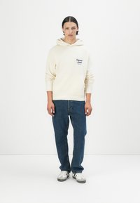 Felpa crema con tasca, testo blu navy, abbinata a jeans blu scuro e sneakers bianche con strisce nere. Semplice, vestibilità rilassata, design minimale.