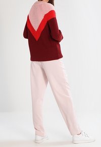 Femme portant un pull en tricot à motifs chevrons rouge, rose et bordeaux avec un pantalon rose clair et des baskets blanches, se tenant les mains dans les poches.