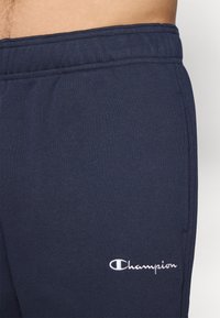 Tamsiai mėlynos treniruotės kelnės, pagamintos iš minkštos medžiagos, su elastinga juosmens juosta ir balta siuvinėta "Champion" logotipu šone.