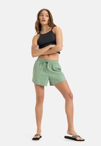 Groene linnen shorts met een elastische tailleband en een trekkoord, gecombineerd met een zwarte geribbelde tanktop en zwarte flip-flops met een wit patroon.