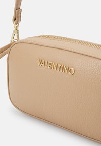 Beige teksturert lær crossover-veske med gullfarget hardware og merkevarelogo. Har glidelås og justerbar stropp.