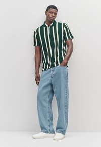 Grün-weiß gestreiftes Poloshirt mit Reißverschluss-Kragen, kombiniert mit hellblauen, locker sitzenden Jeans und weißen Turnschuhen.