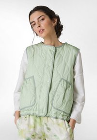 Hellgrüne gesteppte Weste mit rundem Halsausschnitt, Druckknöpfen und zwei Vordertaschen; getragen über einem weißen Langarmshirt.