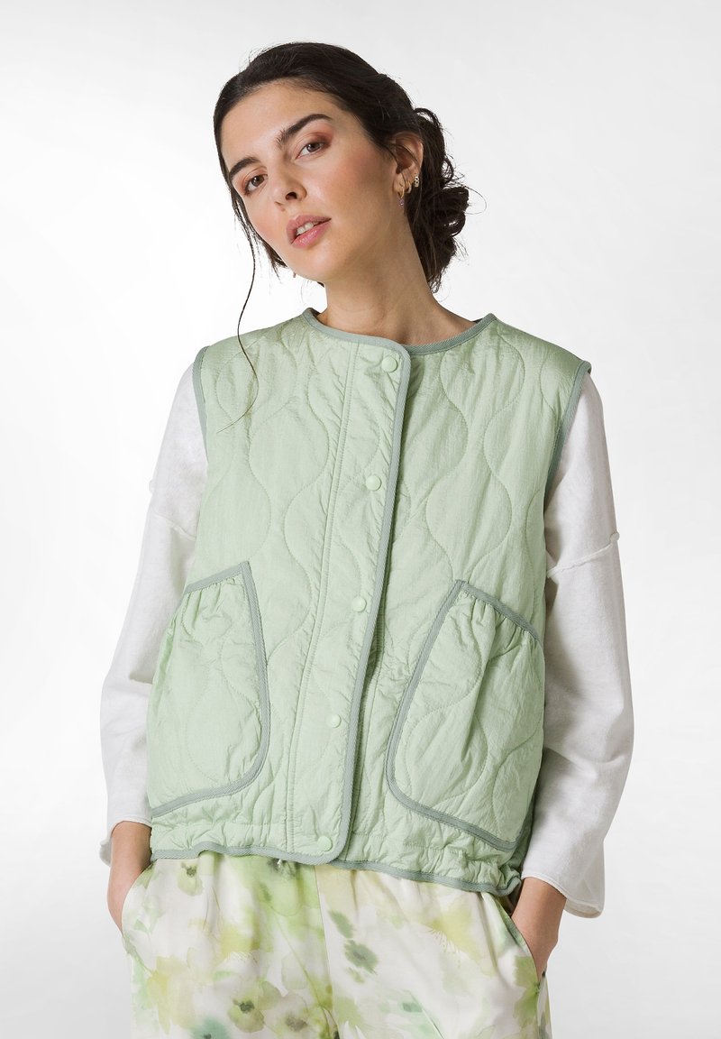 Hellgrüne gesteppte Weste mit rundem Halsausschnitt, Druckknöpfen und zwei Vordertaschen; getragen über einem weißen Langarmshirt.