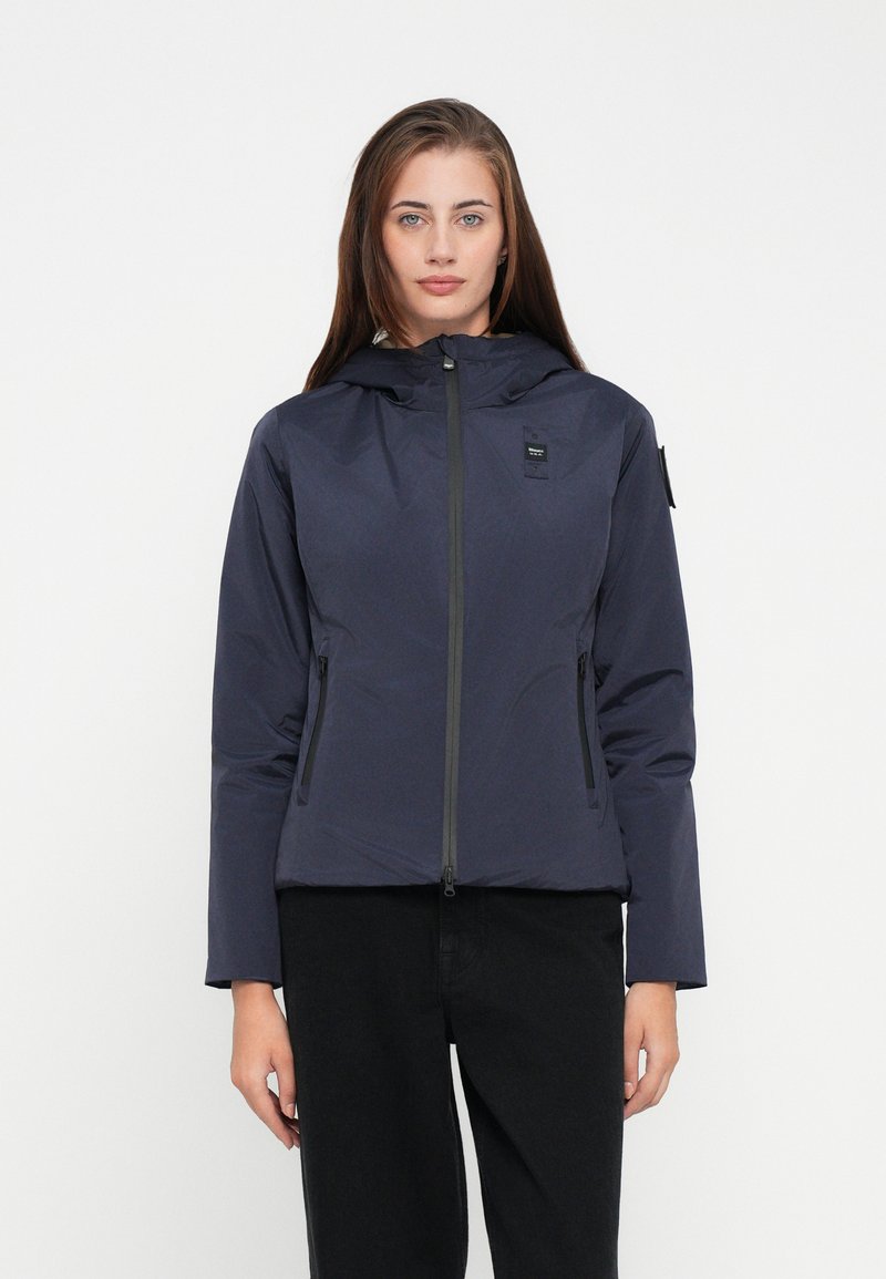 Marineblauwe jack met capuchon, gemaakt van een gladde, lichte stof; voorzien van een zipper aan de voorkant en twee zijzakken. Minimalistisch design met logo.
