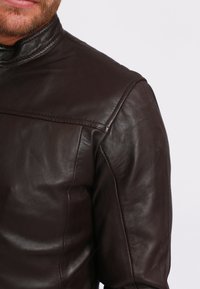 Giacca di pelle marrone con colletto alto, caratterizzata da una texture liscia, linee pulite e un sottile dettaglio di cucitura sulla spalla.