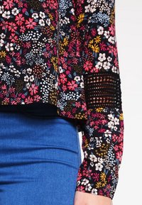 Floral-patterned blouse in black, featuring vibrant pink, white, and yellow flowers. Includes textured accents on the sleeves. Paired with blue jeans.