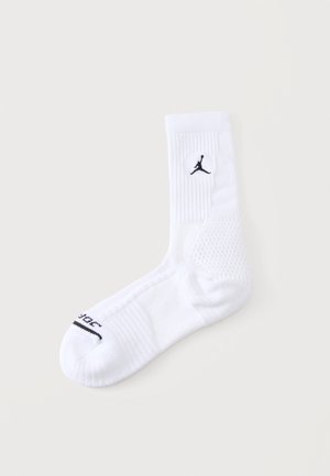 Valge sportlik poolpikk sokk musta Jumpman'i logoga pahkluu piirkonnas ja musta tekstiga varbaosas ühevärvilise tausta ees.