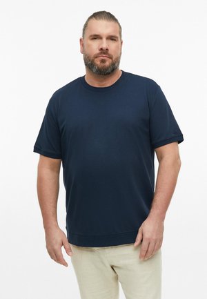 Man met baard en paardenstaart draagt een marineblauw T-shirt met korte mouwen en lichtbeige broek, staande tegen een effen witte achtergrond.