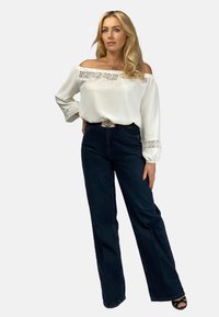 Biała bluzka na jedno ramię z koronkowym wykończeniem przy dekolcie i mankietach, zestawiona z ciemnoniebieskimi szerokimi dżinsami. Casualowy i luźny fason.